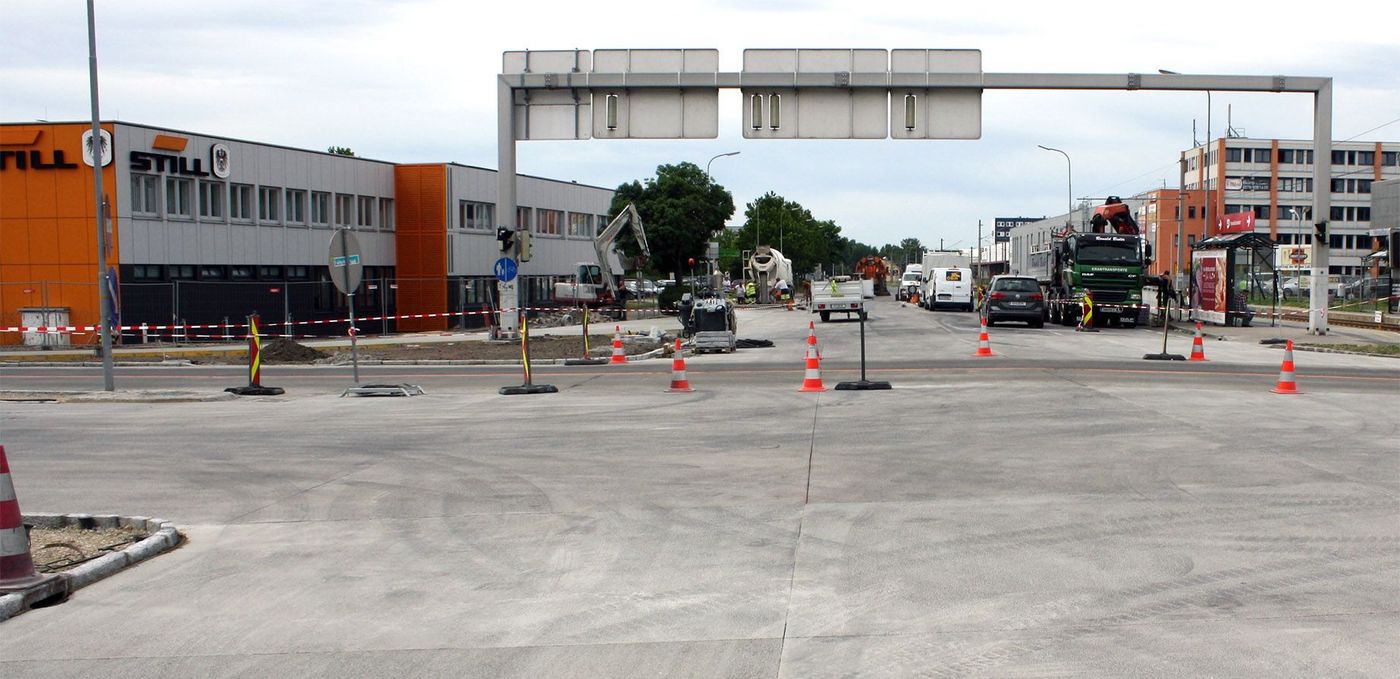 Foto: Frisch betonierte Fläche einer Kreuzung; im Hintergrund Baufahrzeuge, Warnkegel und Gebäude eines Gewerbegebiets