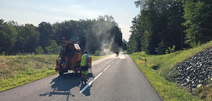 Foto: Asphaltierte Freilandstraße umgeben von Wald- und Wiesenflächen; in der Mitte ein Arbeiter mit Baumaschine, im Hintergrund eine weitere Baumaschine umhüllt von weißem Dampf