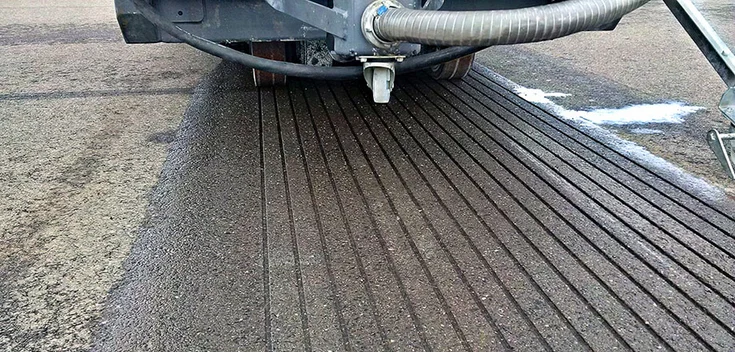 Foto: Nahaufnahme einer Fahrbahnoberfläche, in die Rillen geschnitten wurden; im Hintergrund Teile einer Baumaschine