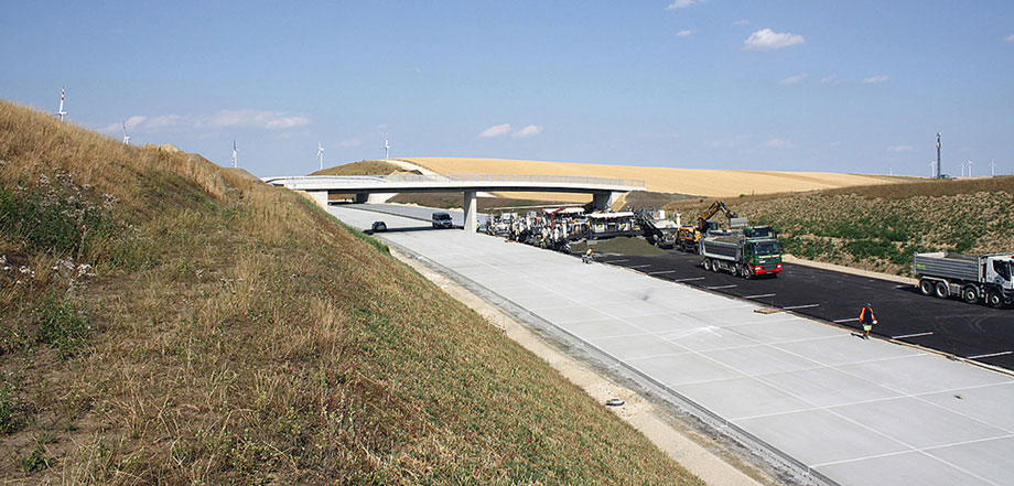 Foto: Frisch betonierter Autobahnabschnitt mit Baufahrzeugen und –maschinen; sanft hügelige Agrarlandschaft im Hintergrund