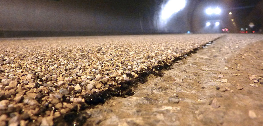 Foto: Nahaufnahme eines grobkörnigen Straßenbelags in einem dunklen Tunnel; im Hintergrund die Scheinwerfer von Baufahrzeugen