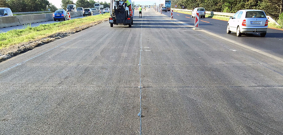 Foto: Neu hergestellter Autobahnabschnitt mit deutlich sichtbaren Fugen, die mit einer schwarzen Masse ausgefüllt sind; im Hintergrund Baufahrzeuge und Arbeiter; rechts und links Fahrzeugverkehr